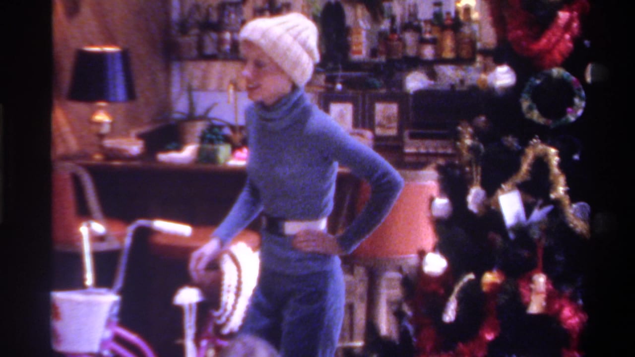 Woman in blue turtleneck and white beanie stands in a Christmas-decorated living room, hand on hip, with a bar visible behind.
