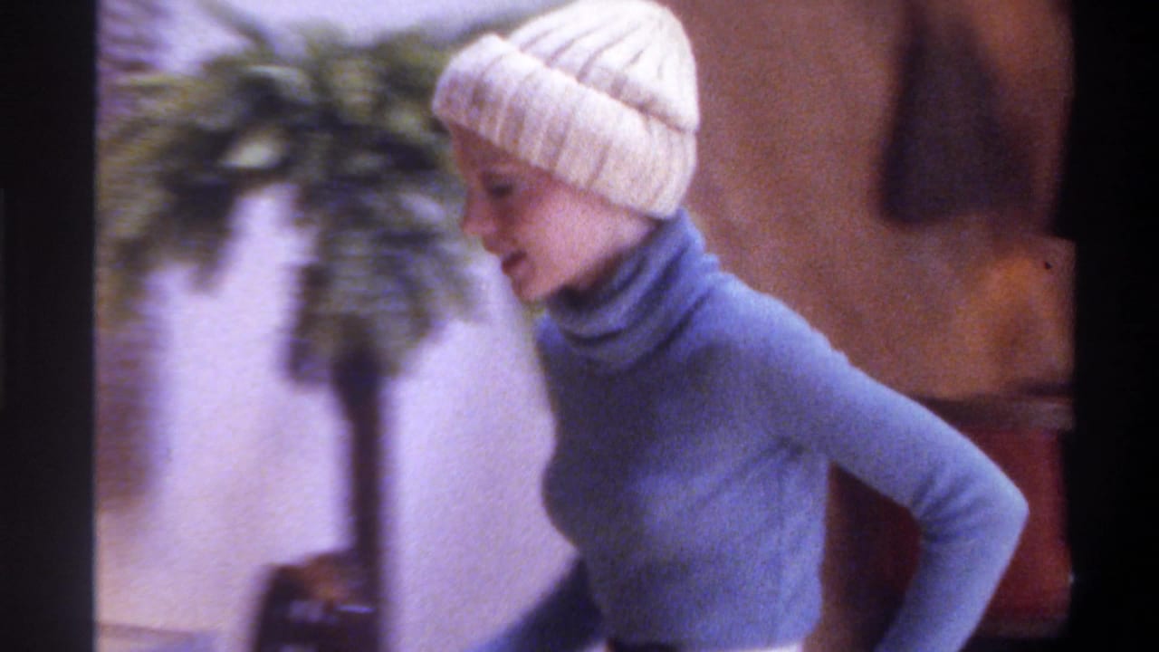 The same woman in a blue turtleneck and white knit beanie pauses beside a large tropical houseplant in a 1970s-style living room interior, likely Gainesville, Florida, 1975.