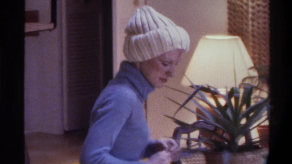 A woman in a blue turtleneck and white knit beanie examines a spiky potted plant beside a floor lamp in a 1970s private living room interior, likely Gainesville, Florida, 1975.