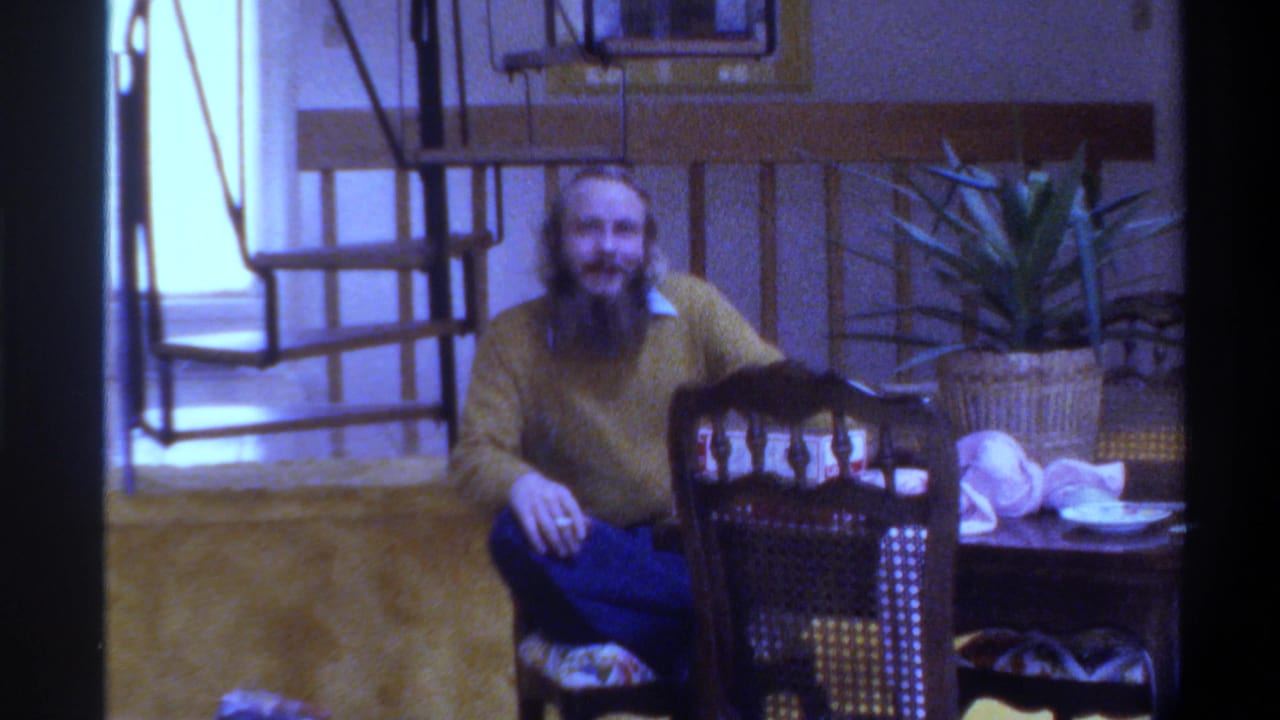 Bearded man in yellow mustard sweater and blue jeans seated at a wooden dining table in a mid-1970s Gainesville, Florida home interior, circa 1975.