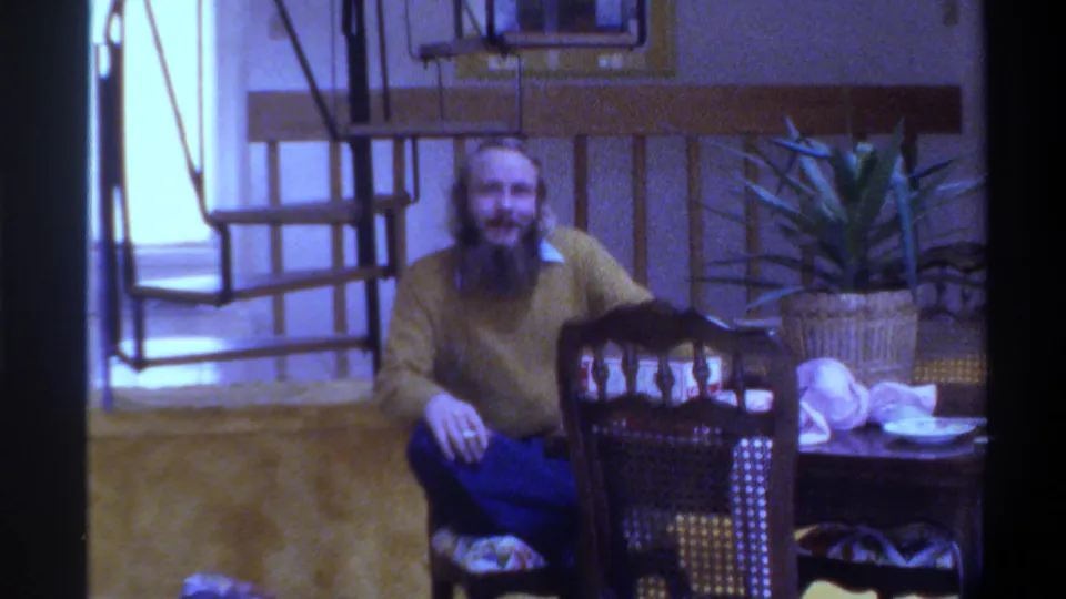 Bearded man in yellow mustard sweater and blue jeans seated at a wooden dining table in a mid-1970s Gainesville, Florida home interior, circa 1975.