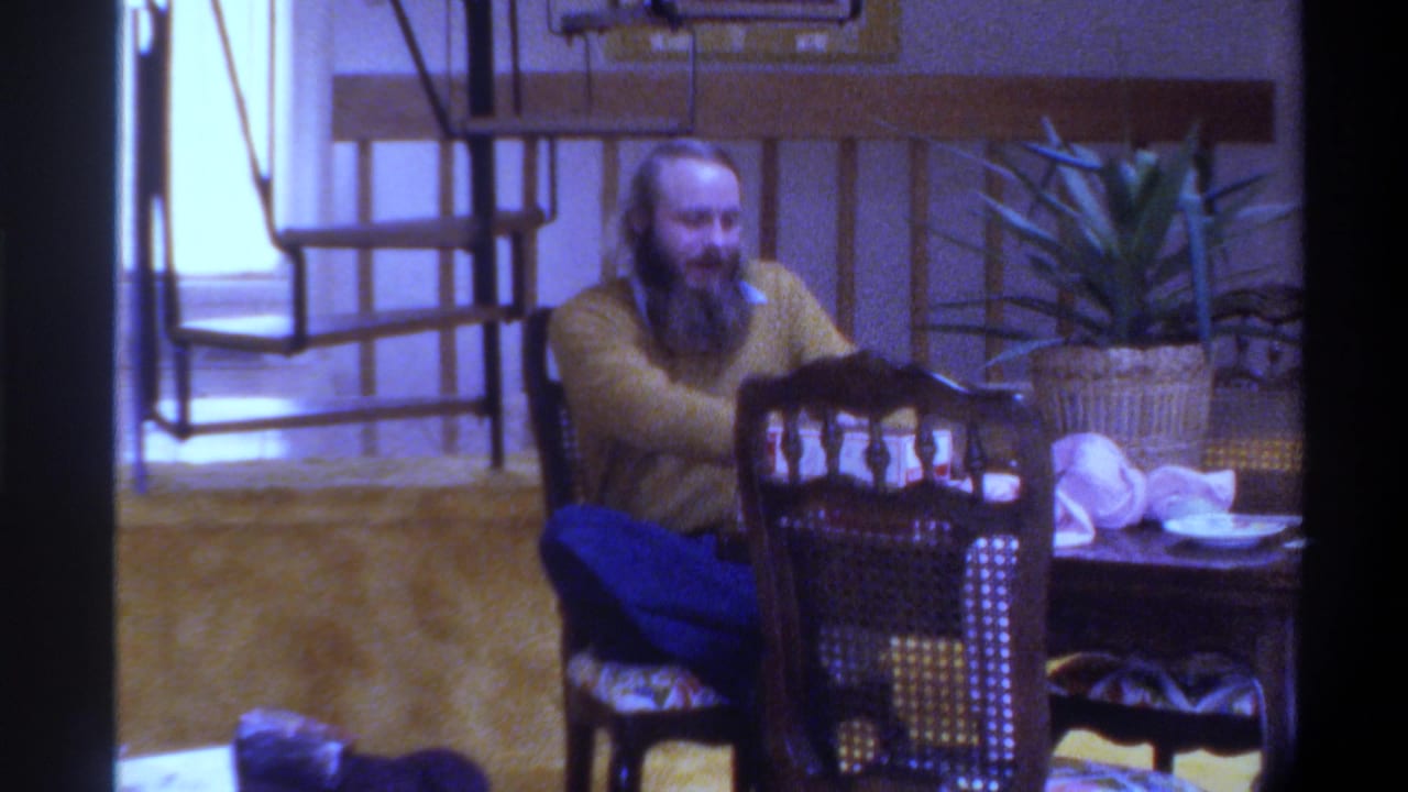 Same bearded man leaning toward table with arm extended, cane-back chairs and potted plant in background.