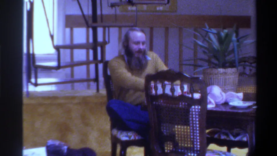 Bearded man in yellow sweater reaching toward dining table in mid-1970s domestic interior, Gainesville, Florida, circa 1975.