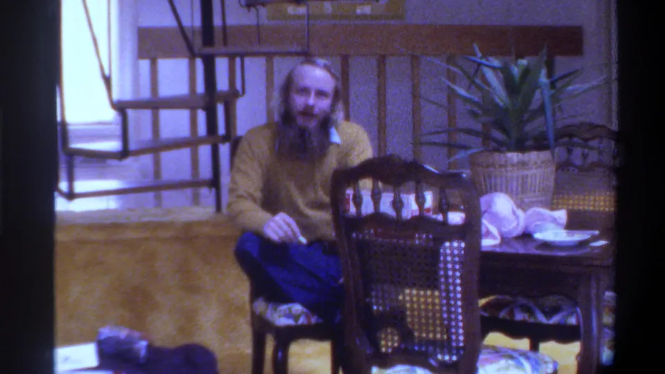 Closer framing of bearded man in yellow sweater at dining table looking toward camera, mid-1970s Gainesville, Florida home interior.