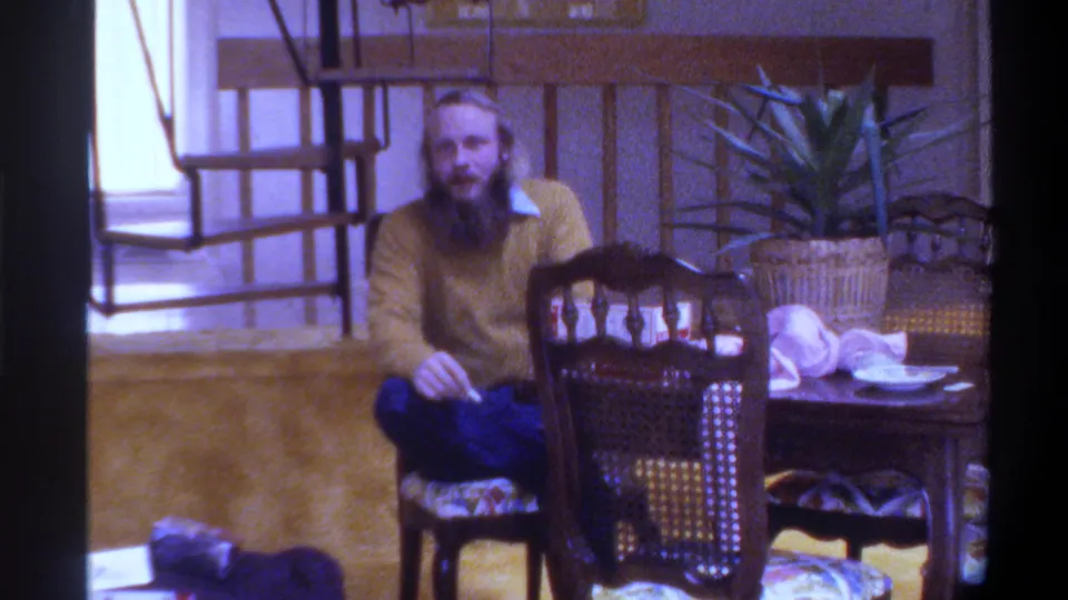 Final frame: bearded man in yellow sweater and blue jeans at dining table in 1975 Gainesville, Florida home interior, looking toward camera.