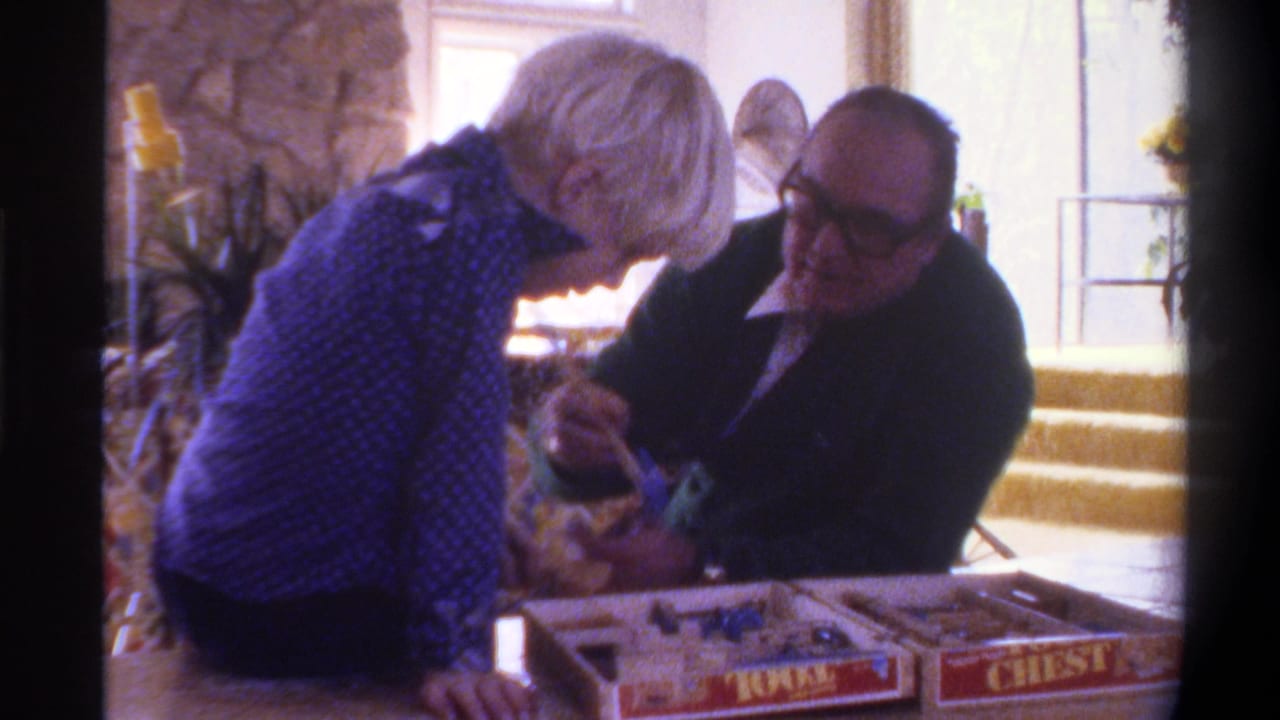 In a 1975 8mm home movie, an elderly man in dark jacket and tie and a light-haired child in a blue patterned shirt focus together on a game set with visible branded red packaging.