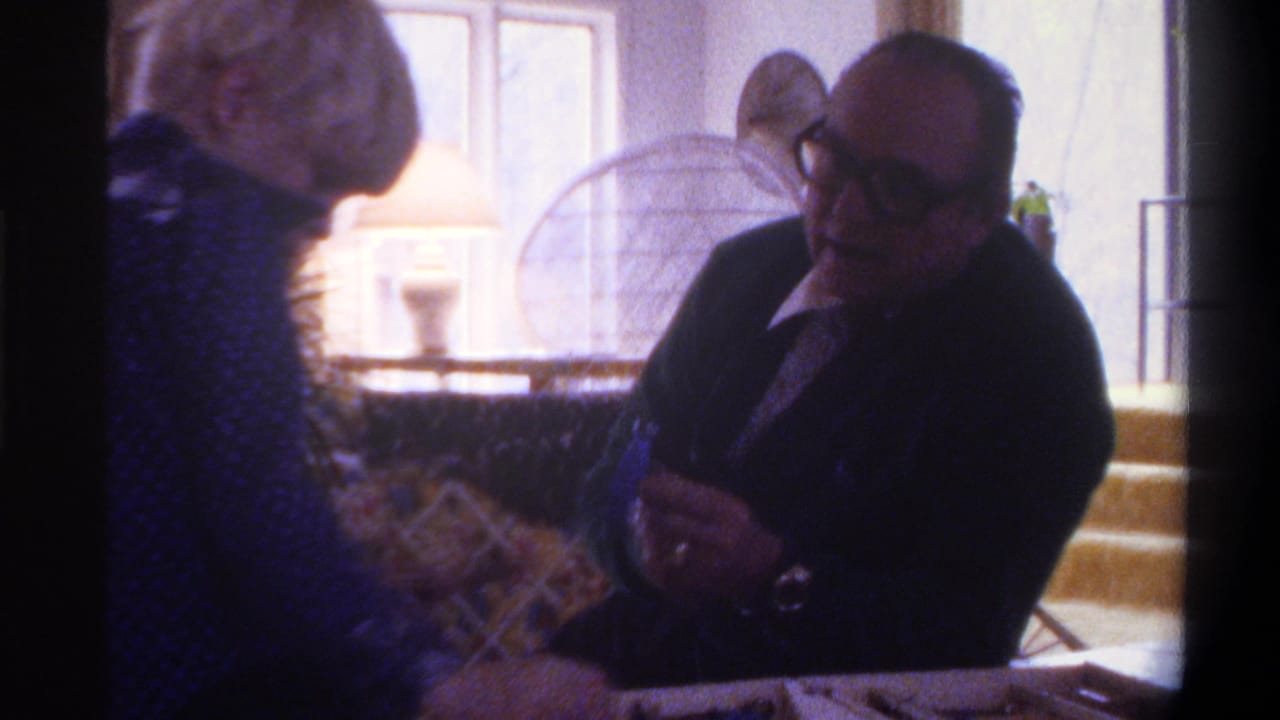 Elderly man with dark-framed glasses in a dark jacket seated at a table with game materials; second figure partially visible at left, stone wall and wire-frame chair behind.