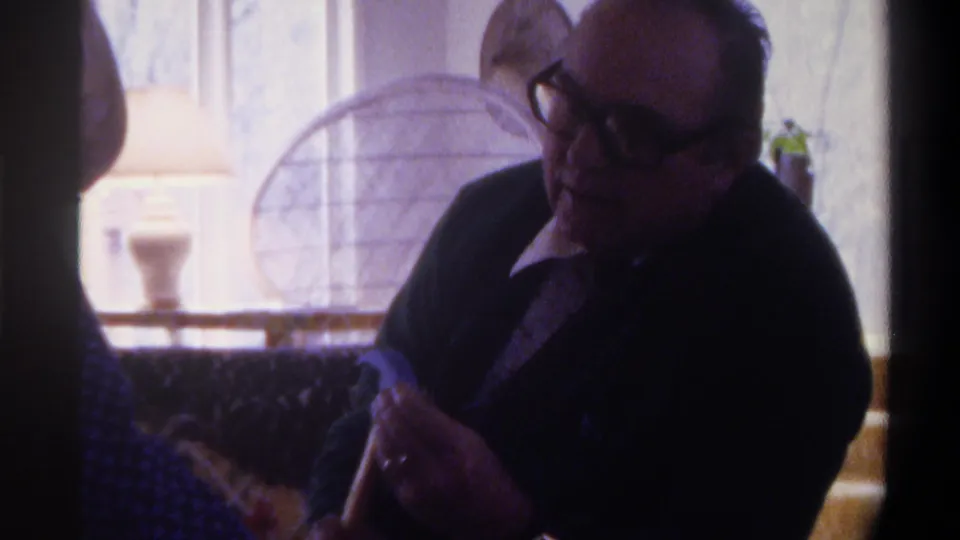 Close medium shot from a 1975 8mm clip; elderly man with large dark-framed glasses and dark jacket looks down, stone-veneer wall and circular wire-frame decoration visible behind him.