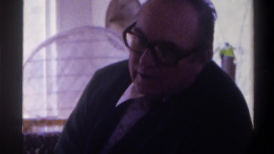 Close shot from a 1975 8mm home movie; elderly man with dark-framed glasses and dark jacket looks to one side in a private living room with stone wall, circular wire element, and plant material.