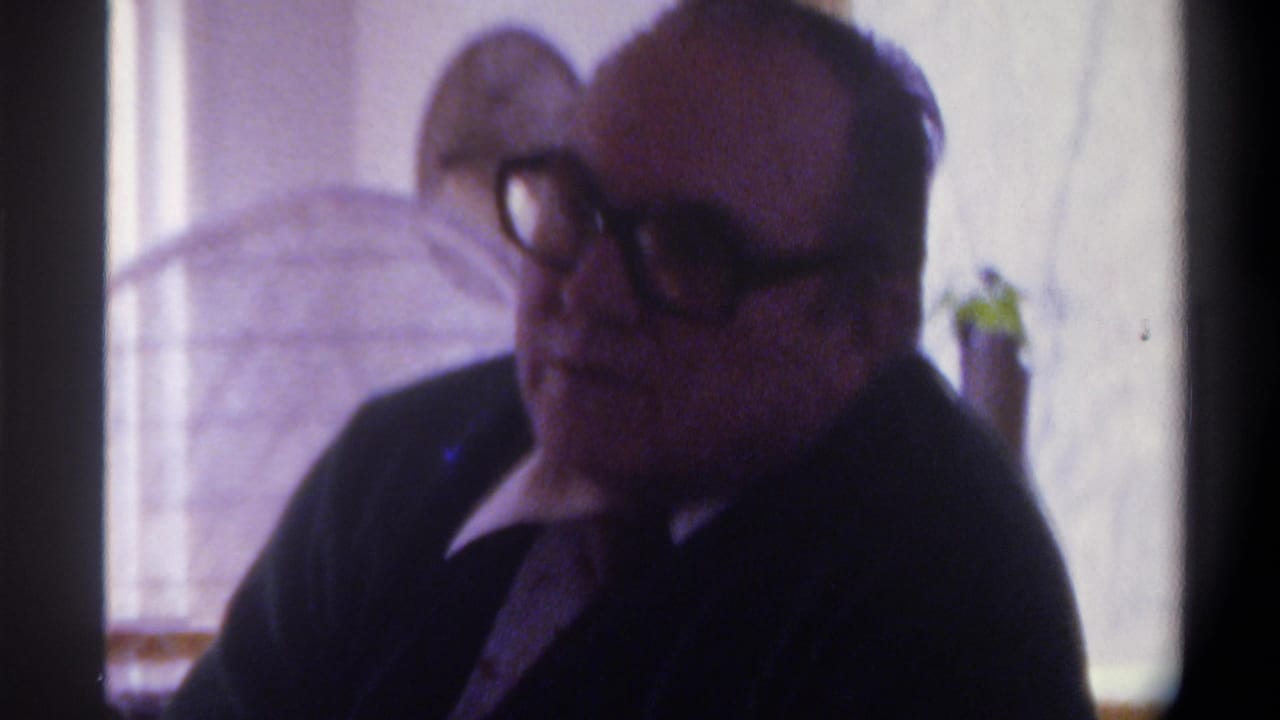 Close-up of a heavyset man with thick dark-framed glasses and thinning hair, wearing a dark suit, facing right; arched window and small plant softly visible in background.