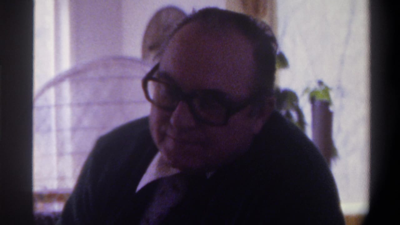 8mm close-up of a heavyset man in thick-framed glasses and dark suit, mid-conversation, Gainesville Florida c.1975; arched window and potted plant in soft-focus background.