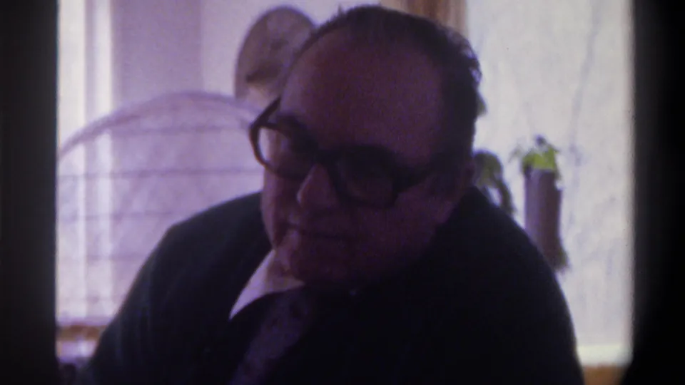 8mm home movie frame of a heavyset man in thick plastic-framed glasses and dark suit, eyes slightly downcast, Gainesville Florida c.1975; indoor window and plant behind him.