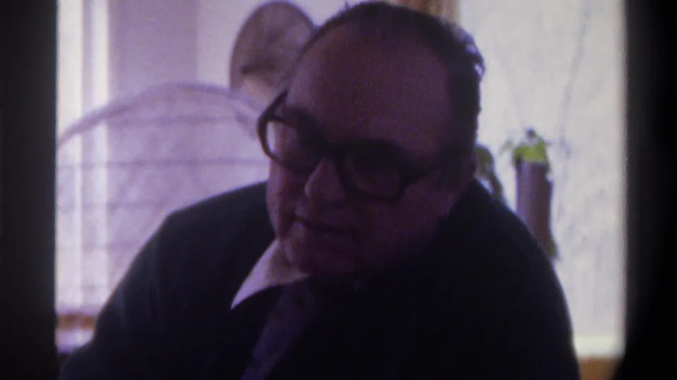 8mm close-up of a heavyset balding man in thick-framed glasses and dark suit, Gainesville Florida 1975; arched window with soft indoor light and plant in background.