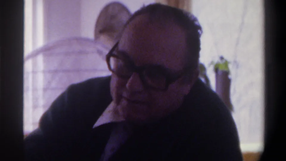 In a 1975 Gainesville, Florida home-movie frame, an older bald man wearing thick dark glasses and a dark cardigan sits in soft indoor light near a bright window.