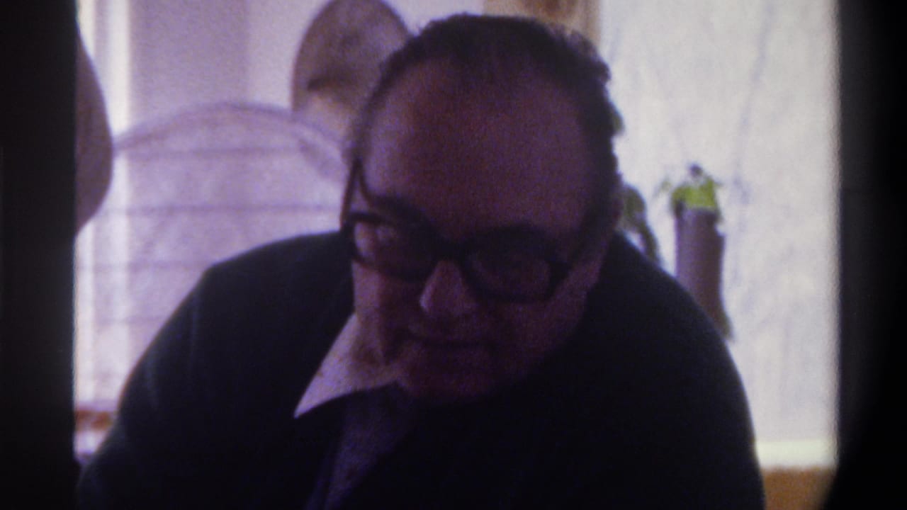 Older man in glasses leans slightly forward in a dim indoor room.