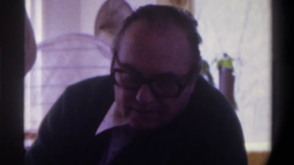 The older man remains seated indoors in the 1975 Gainesville home-movie scene, partly silhouetted against a bright window with soft focus and purple-tinted 8mm color.