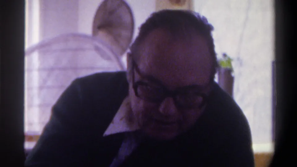 A close 8mm frame shows the older man with dark glasses looking downward in the indoor Gainesville, Florida home-movie setting, with a bright window and household objects behind him.