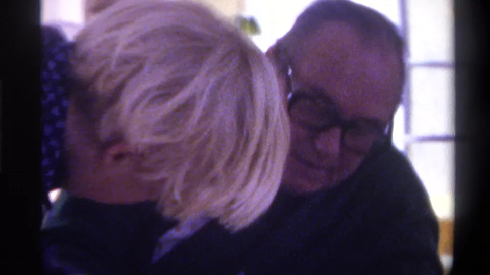 A young blond person leans into the frame near the seated older man with glasses, partially obscuring him during a close indoor conversation in the 1975 Gainesville home movie.