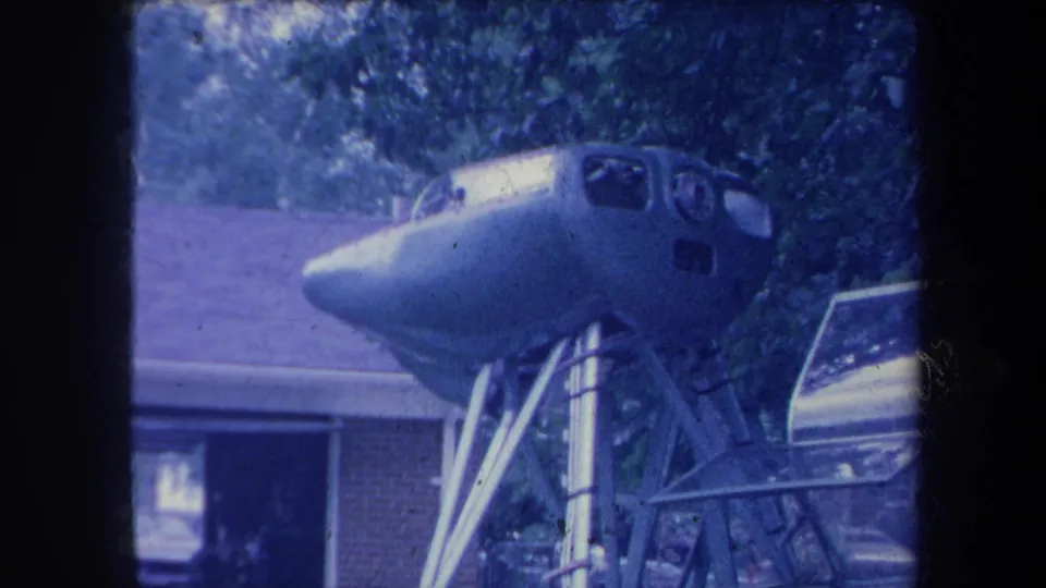 Aircraft nose/cockpit section elevated on a metal stand in a Yucaipa, California driveway, circa 1959, blue-shifted aged 8mm film.