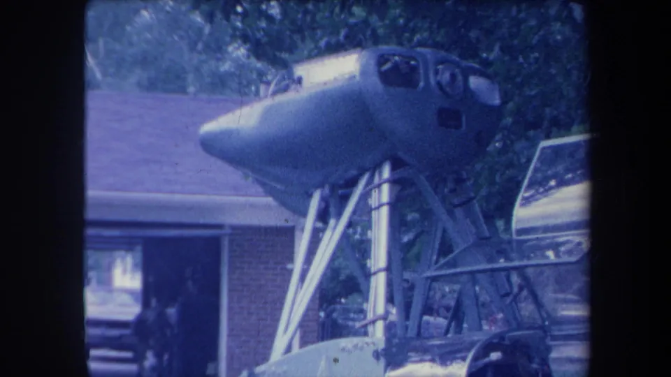 Aircraft cockpit nose on tripod stand in driveway with figure near garage doorway, Yucaipa California ca. 1959, aged 8mm film.