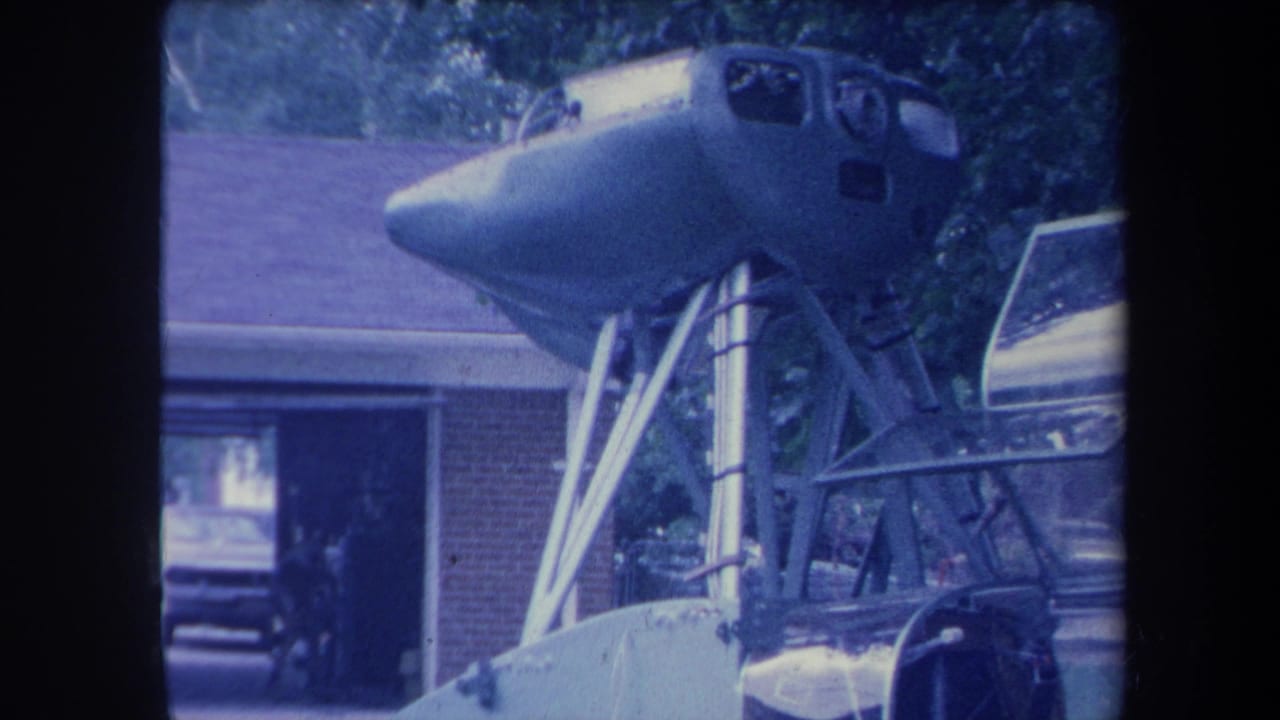 8mm home-movie frame circa 1959 showing a jet aircraft cockpit nose section elevated on a homemade metal support frame, brick ranch house and open garage visible, Yucaipa California area.