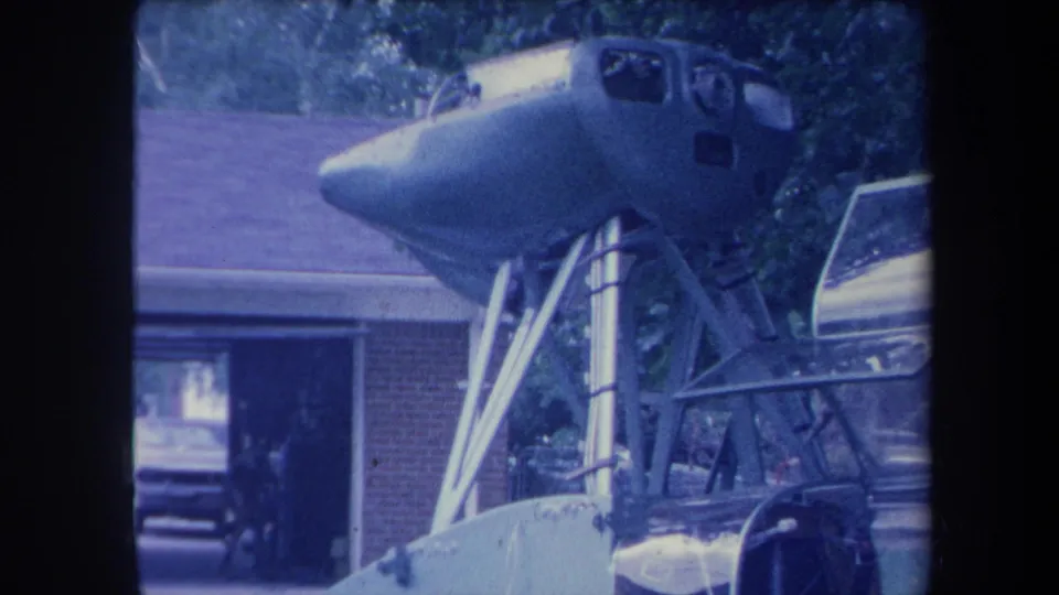 8mm home-movie frame circa 1959 showing a jet aircraft cockpit nose section elevated on a homemade metal support frame, brick ranch house and open garage visible, Yucaipa California area.