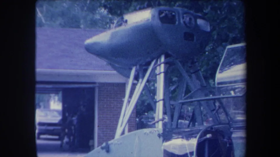 8mm frame circa 1959 showing a jet aircraft nose and cockpit section elevated on metal struts beside a brick suburban house, a vintage car visible in the open garage, Yucaipa California area.