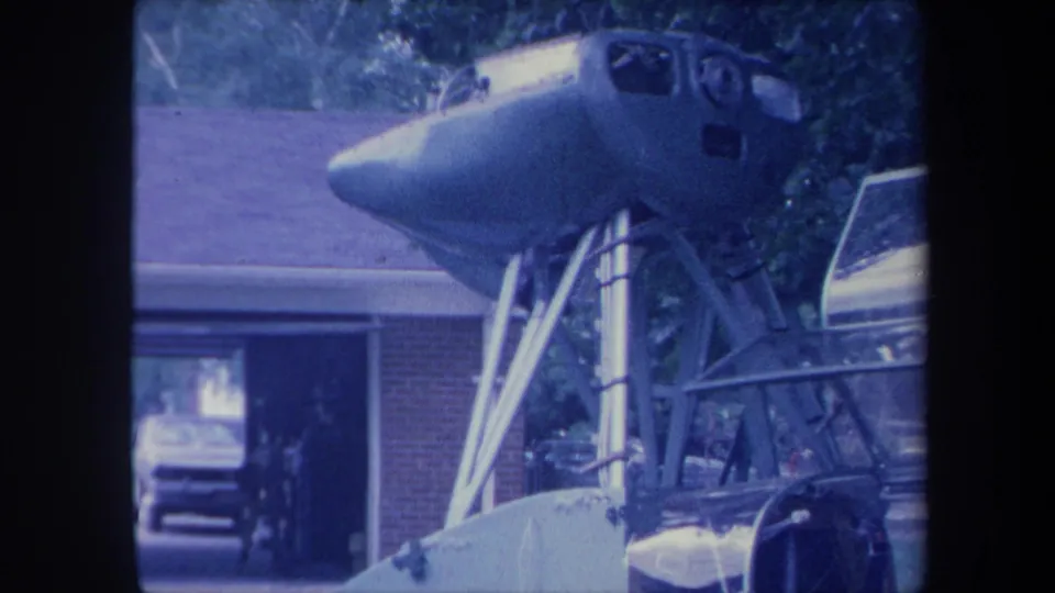 8mm frame circa 1959 revealing more of the lower metal frame construction beneath a jet cockpit section, brick ranch house and partial vehicle visible in background, Yucaipa California area.