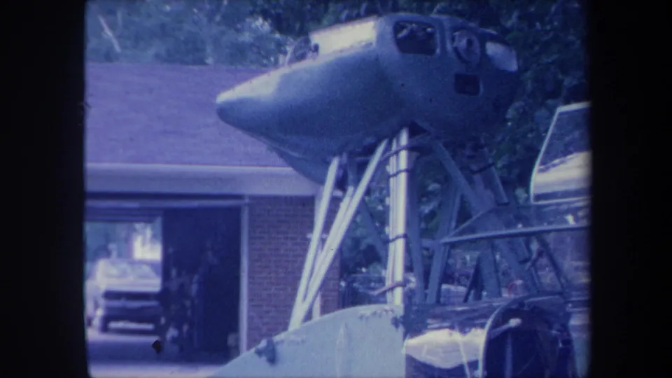 Final 8mm frame circa 1959 showing the jet cockpit nose section angled forward on its support frame, brick suburban house roofline and partial vintage automobile visible at right, Yucaipa California area.