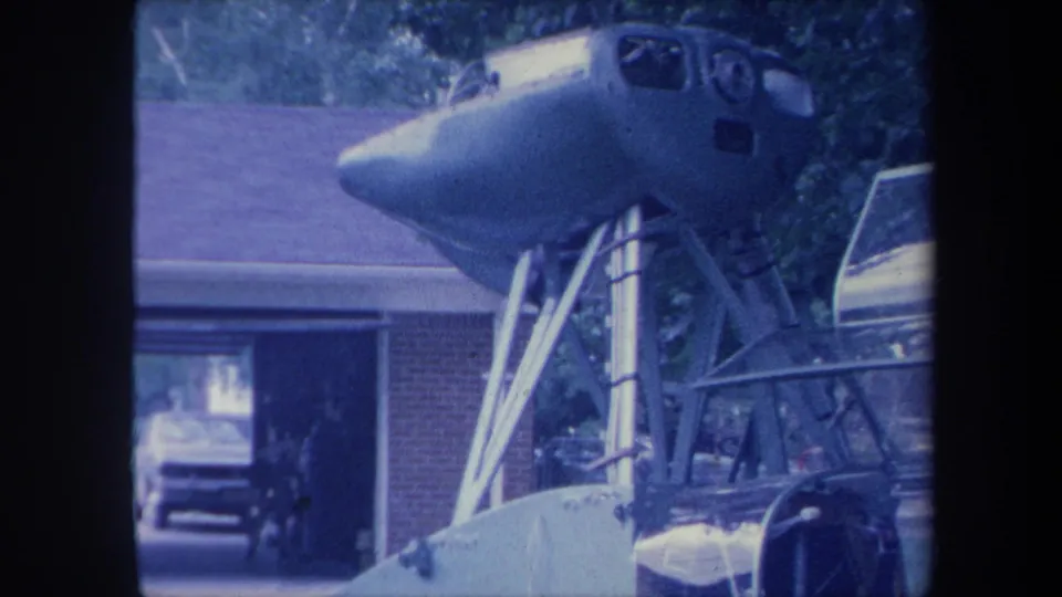 Homemade aircraft fuselage on a metal scaffold in a residential driveway, Yucaipa California circa 1959, filmed in 8mm with blue-purple color shift.
