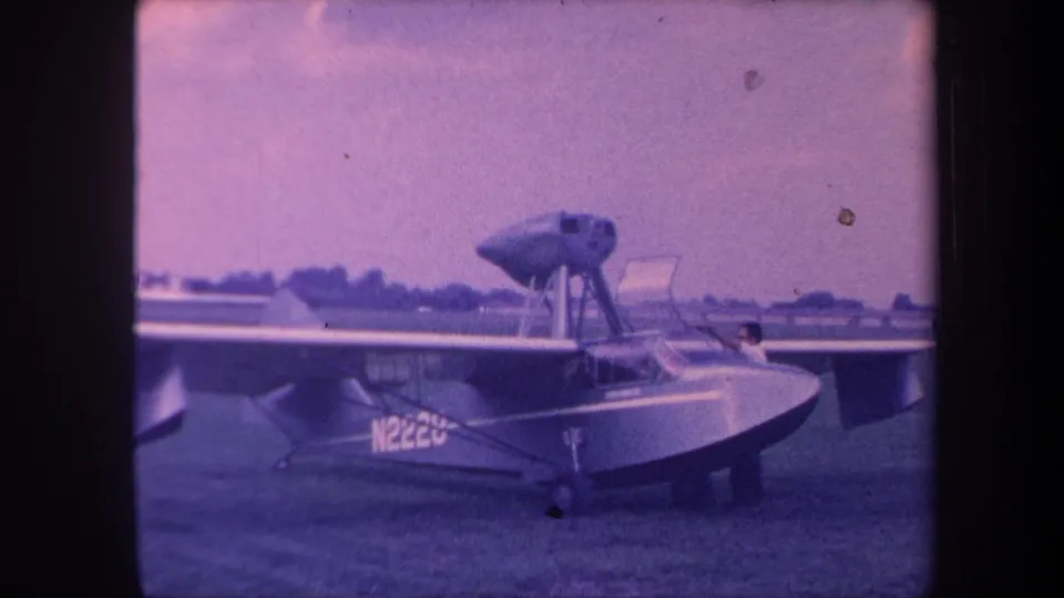8mm exterior of a small homebuilt amphibious aircraft — boat hull fuselage, bubble canopy, wing-mounted floats, partially readable N-number — on a California airfield circa 1959; figure visible near cockpit.