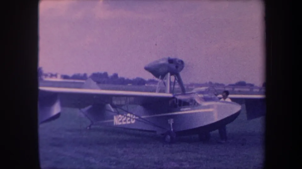 8mm home movie still, circa 1959, Yucaipa California airfield; amphibious aircraft N2220 with occupant visible in cockpit area on grassy field.