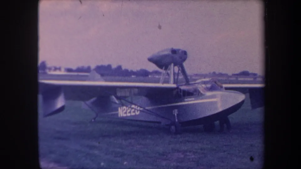 Amphibious flying boat N222U on grass airfield, California circa 1959; this frame offers a slightly closer view of the boat hull and propeller nacelle.