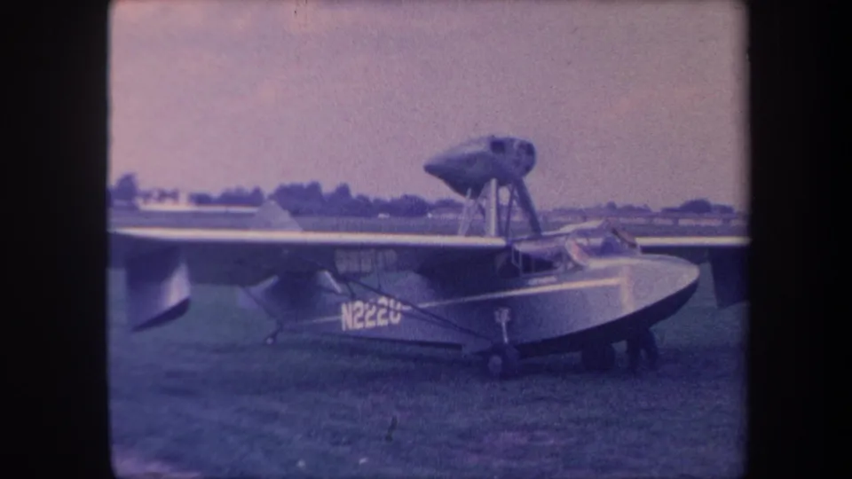 1950s amphibious aircraft on a California grass airfield, boat-hull fuselage and strut-mounted engine visible, filmed on 8mm home movie stock.