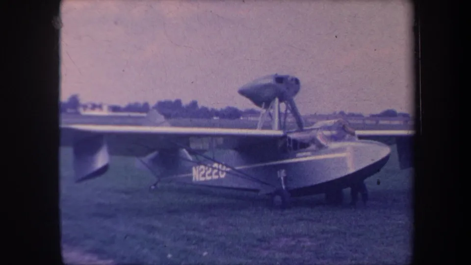 High-wing amphibious aircraft with boat hull on a grass airfield, engine struts and partially legible registration visible, 8mm color shift, circa 1959 California.