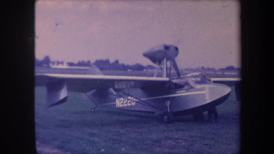 Ground-level view of 1950s amphibious aircraft hull and keel on grassy California airfield, registration partially legible, blue-shifted 8mm stock.