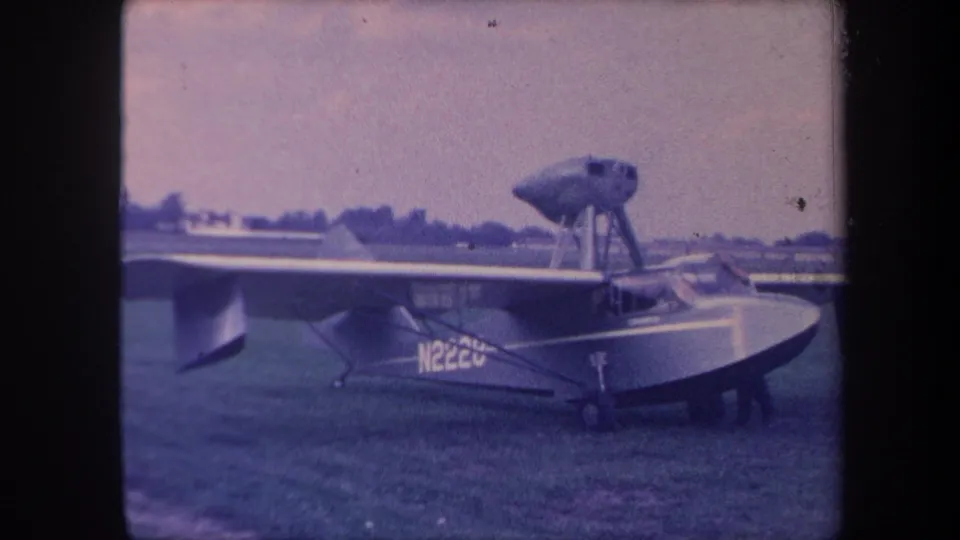 Final frame of 1950s amphibious aircraft sequence on California grass strip, high-wing silhouette and boat hull against overcast, 8mm grain throughout.