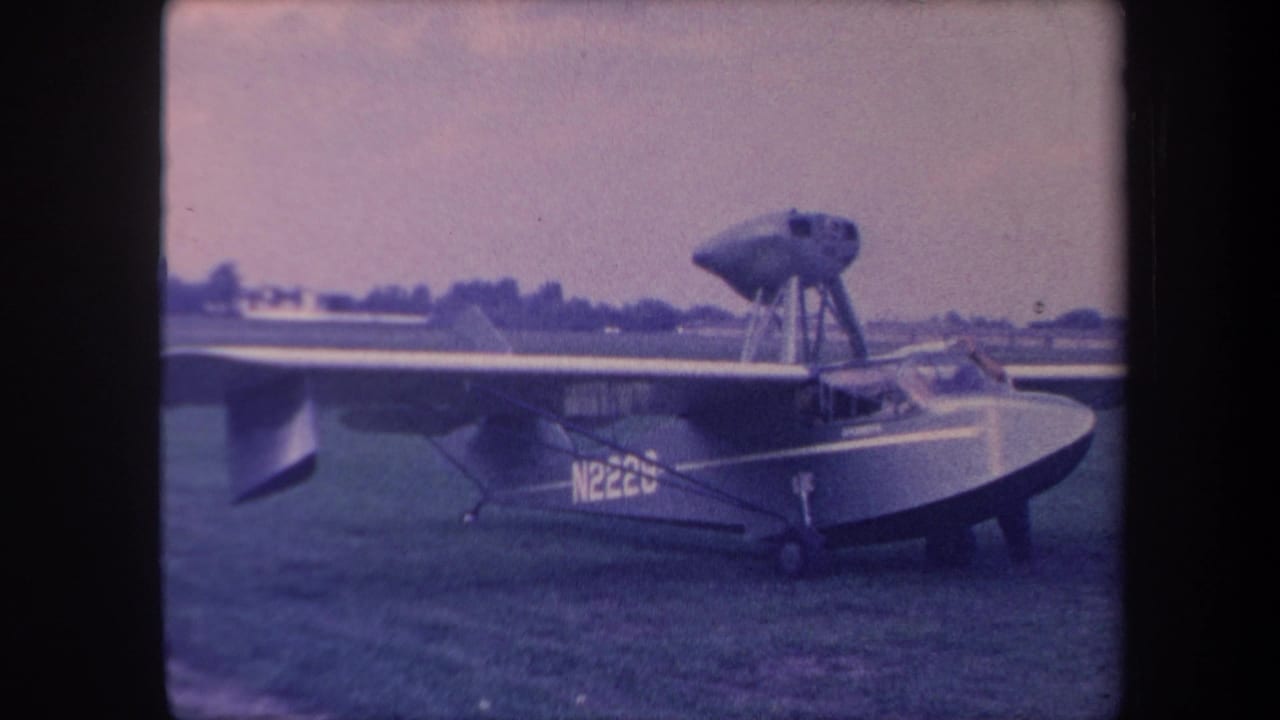 Same amphibious aircraft N222U on grass, slightly wider framing showing right wing, high pusher engine on strut pylon visible.
