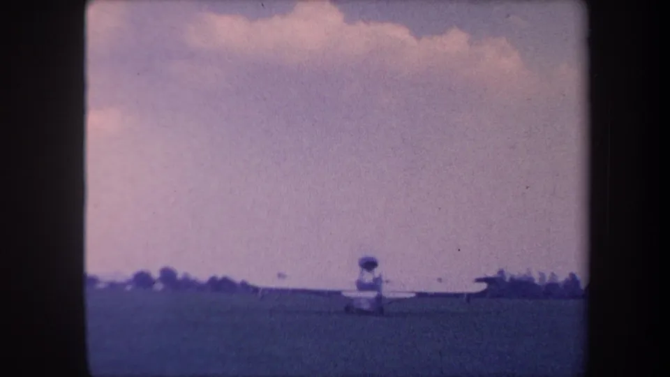 8mm home movie, c.1959, wide pull-back shot of grass airfield with amphibious aircraft in background and unidentifiable human figure in midground.