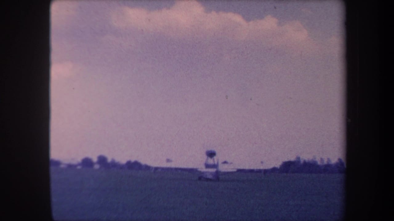 Small aircraft near a tower structure at the horizon of a flat grass airfield, pale purple-blue sky above, 1959 8mm home movie.