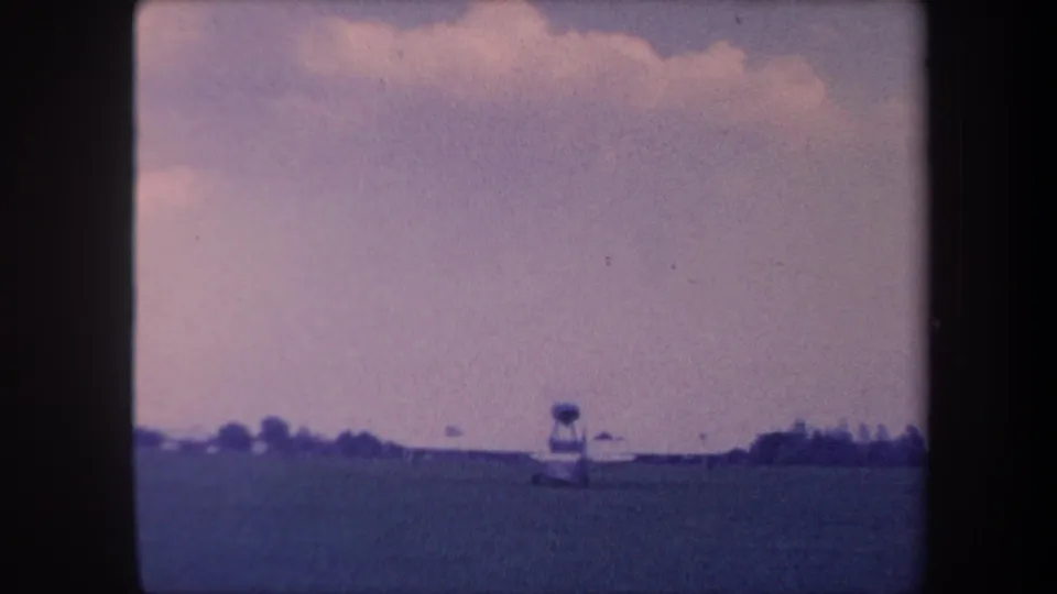 Small propeller aircraft on a grass airstrip near Yucaipa, California, circa 1959, with a vertical structure and distant tree line under a purple-tinged cloudy sky.