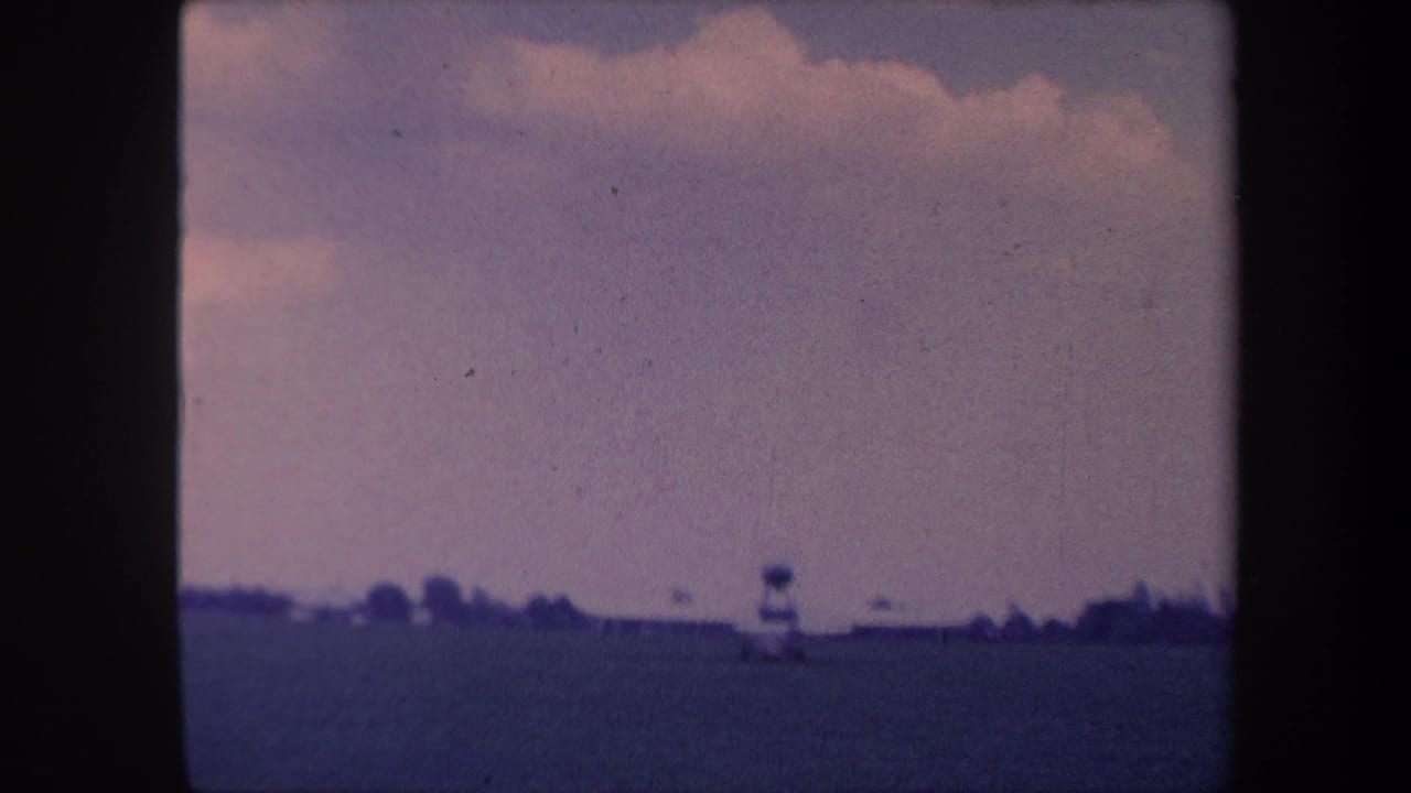 Small aircraft taxiing across a flat grass field, tower structure visible at horizon, pale cloudy sky above.