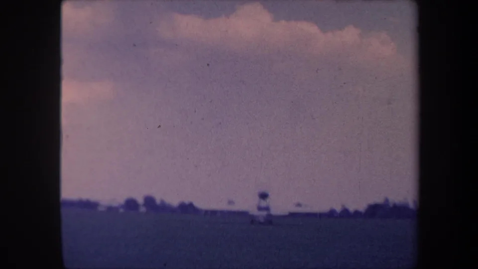 Small propeller aircraft moving left across a grass airstrip near Yucaipa, California, circa 1959, with cumulus clouds and a low tree line at the horizon.