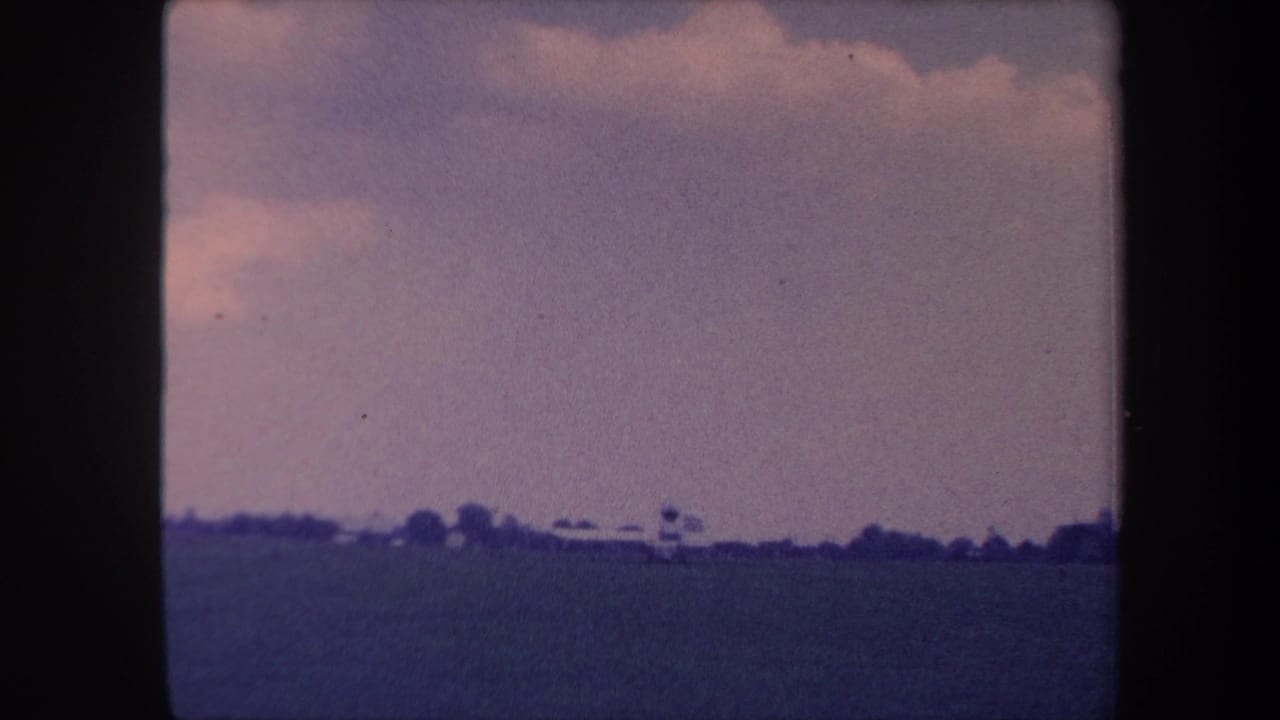 1959 8mm home movie frame near Yucaipa, California, with a wide field, distant treeline, possible windmill or tower structure, and a faded blue-purple sky.