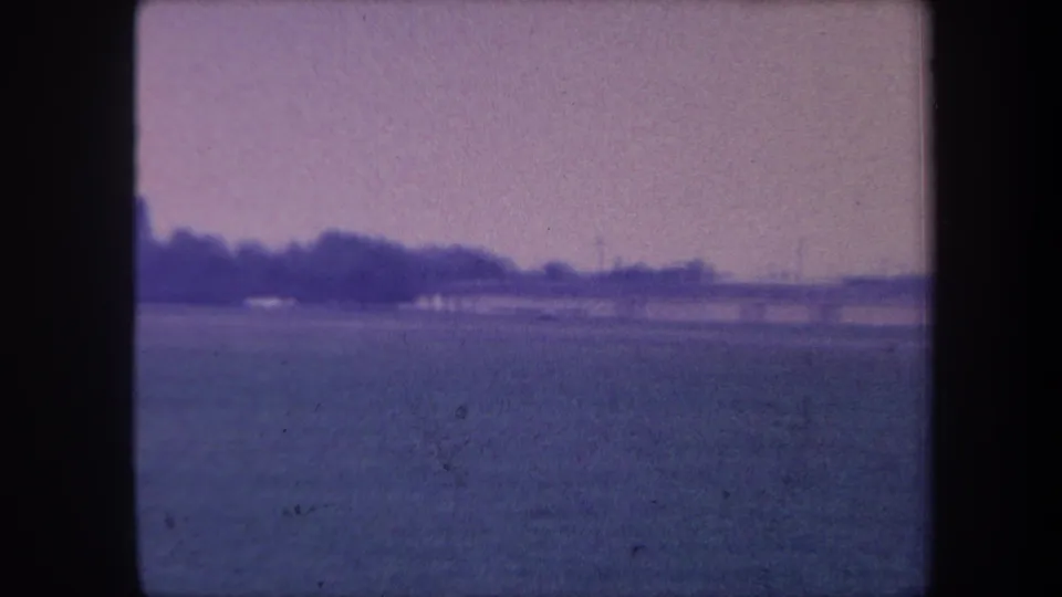 1959 8mm home movie frame near Yucaipa, California — wide foreground field, a dark treeline at mid-distance, and a pale lavender sky with slight blur.