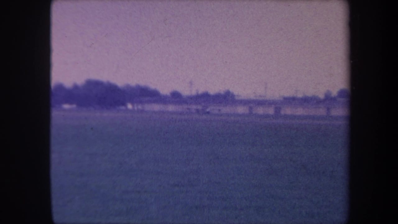8mm home movie frame, circa 1959, showing a flat rural airstrip in Yucaipa, California: open grass field, distant treeline, and blue-purple dye-shifted sky.