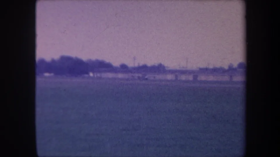 8mm home movie frame, circa 1959, showing a flat rural airstrip in Yucaipa, California: open grass field, distant treeline, and blue-purple dye-shifted sky.