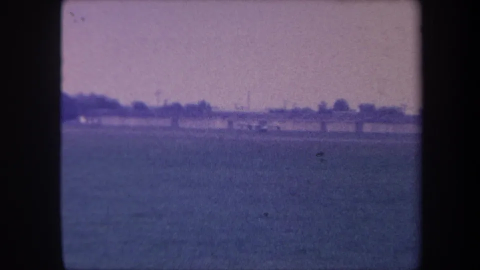 8mm frame circa 1959, Yucaipa, California: flat grassy airstrip with muted purple sky, horizon treeline, and indistinct low structures in the distance.