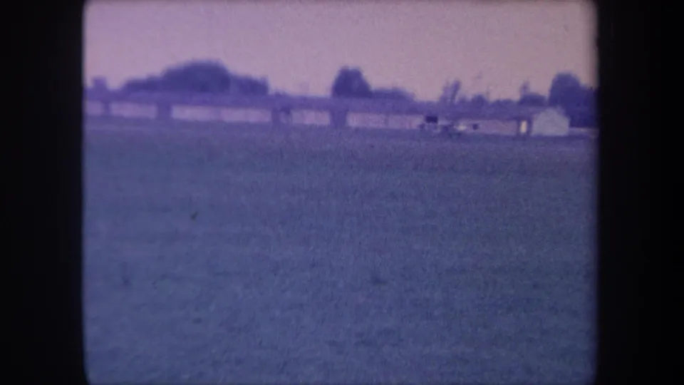 8mm home movie frame, circa 1959, Yucaipa, California; small aircraft near low hangar buildings at the right edge of a grass airfield.