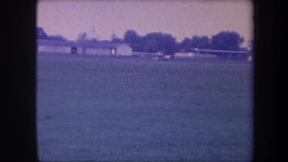 8mm home movie frame, circa 1959, Southern California; small propeller aircraft on the runway with low buildings and a treeline behind.